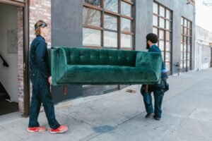 Two removal specialists carrying an old green sofa outdoors during a sofa removal Dubai collection